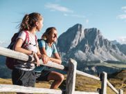 Two female hikers lean on a wooden fence enjoying the view of striking mountain peaks in summer.
