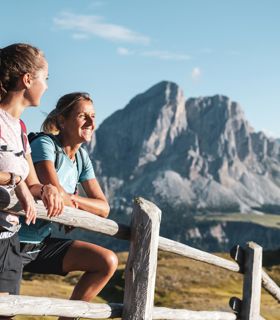 Two female hikers lean on a wooden fence enjoying the view of striking mountain peaks in summer.