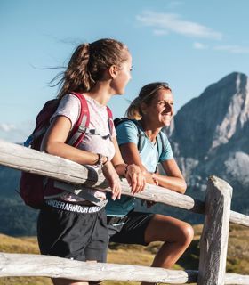 Two female hikers lean on a wooden fence enjoying the view of striking mountain peaks in summer.