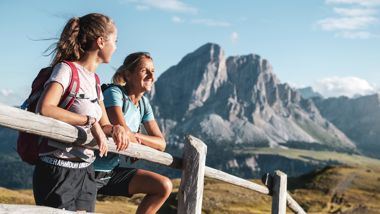 Two female hikers lean on a wooden fence enjoying the view of striking mountain peaks in summer.