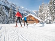 Two cross-country skiers on a snowy trail passing a wooden hut in a winter mountain landscape.