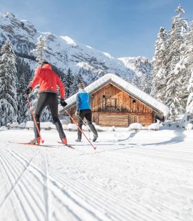 Two cross-country skiers on a snowy trail passing a wooden hut in a winter mountain landscape.