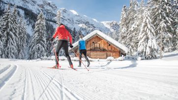Due sciatori di fondo su una pista innevata davanti a una baita alpina e un bosco invernale.