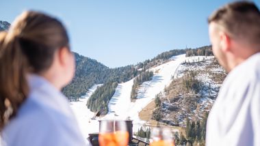 Couple in bathrobes with aperitif watches the sunny ski slope and forested mountain slopes.