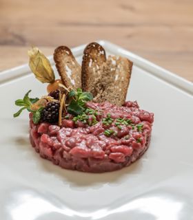 Rindertatar auf weißem Teller, garniert mit Kräutern, Beeren, Physalis und knusprigem Brot.