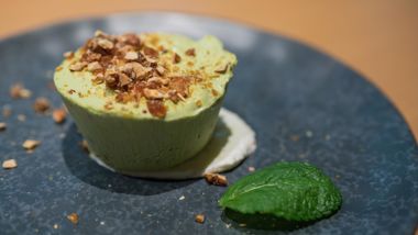 Pistachio parfait on dark plate, topped with brittle, mint leaf and a dollop of cream.