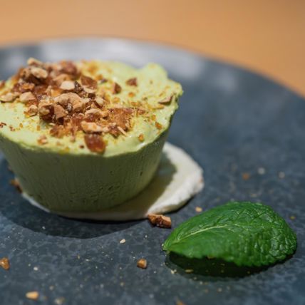 Pistazienparfait auf dunklem Teller, garniert mit Krokant, Minzblatt und Sahnetupfer.