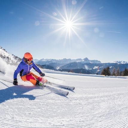 Two skiers carving on fresh piste under bright sun with stunning mountain scenery.