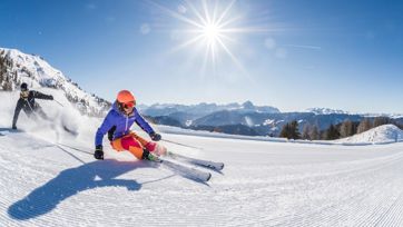 Two skiers carving on fresh piste under bright sun with stunning mountain scenery.