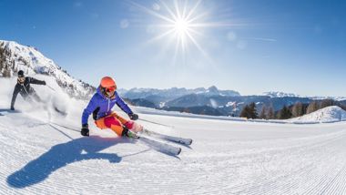 Two skiers carving on fresh piste under bright sun with stunning mountain scenery.