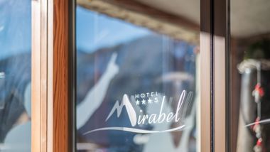 Glass front with Hotel Mirabel logo; mountains and blue sky reflected in the background.