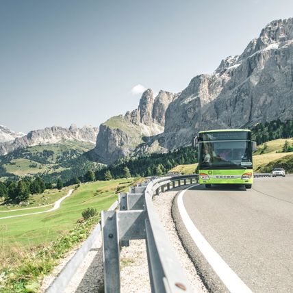 Public bus on a mountain road, surrounded by green meadows and the rocky Dolomites.