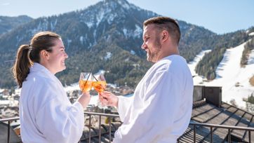 Couple in bathrobes toasting with Aperol, snowy mountains in the background.