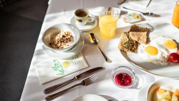 Hearty breakfast with eggs, cheese, ham, muesli, juice, croissant, and fruit on a set table.