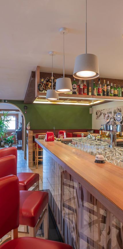Unsere gemütliche Hotelbar mit roten Polsterstühlen, grün gestrichenen Wänden und gut bestücktem Spirituosenregal hinter dem Tresen, beleuchtet von modernen Hängelampen.