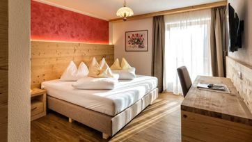 Gemütliches Doppelzimmer mit Holzmöbeln, rotem Akzent, Schreibtisch, TV und Balkonzugang.