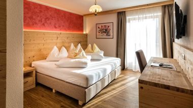 Gemütliches Doppelzimmer mit Holzmöbeln, rotem Akzent, Schreibtisch, TV und Balkonzugang.